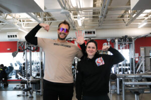 image of fitness equipment and mirrors in the background with smiling Gordie Gronkowskie wearing a tan tee, black pants, and multi-colored glasses flexing his muscles with a smiling Jesse Labreck, wearing a black hoodie and black pants.