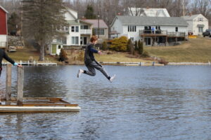 A young boy dressed in long sleeves, pants and white socks leaps into the water from the edge of the dock. 