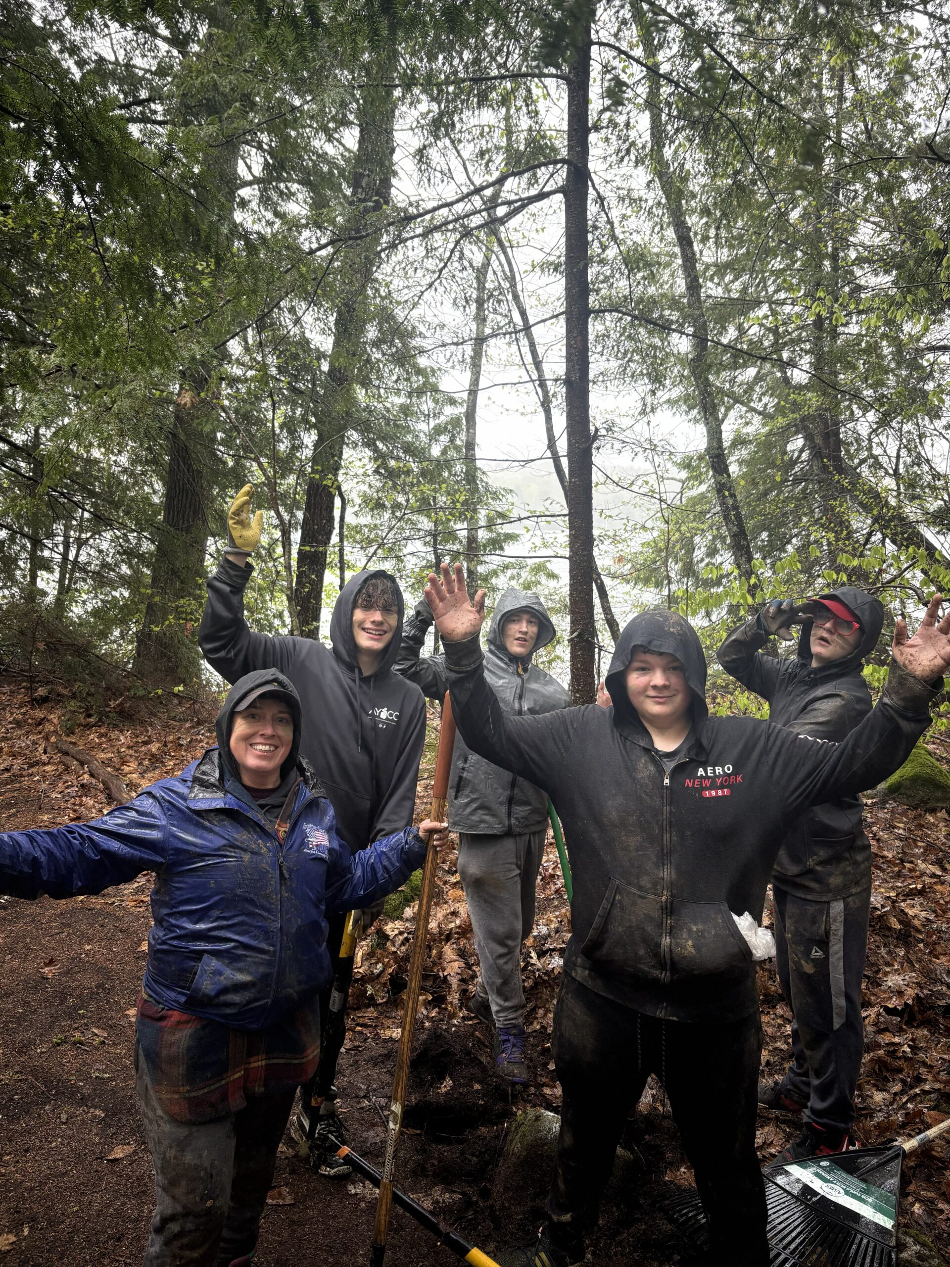 staff and teens standing in the woods, surrounded by trees, and bundled up with hoodies and rain jackets raise their hands in the air in a welcoming manner.