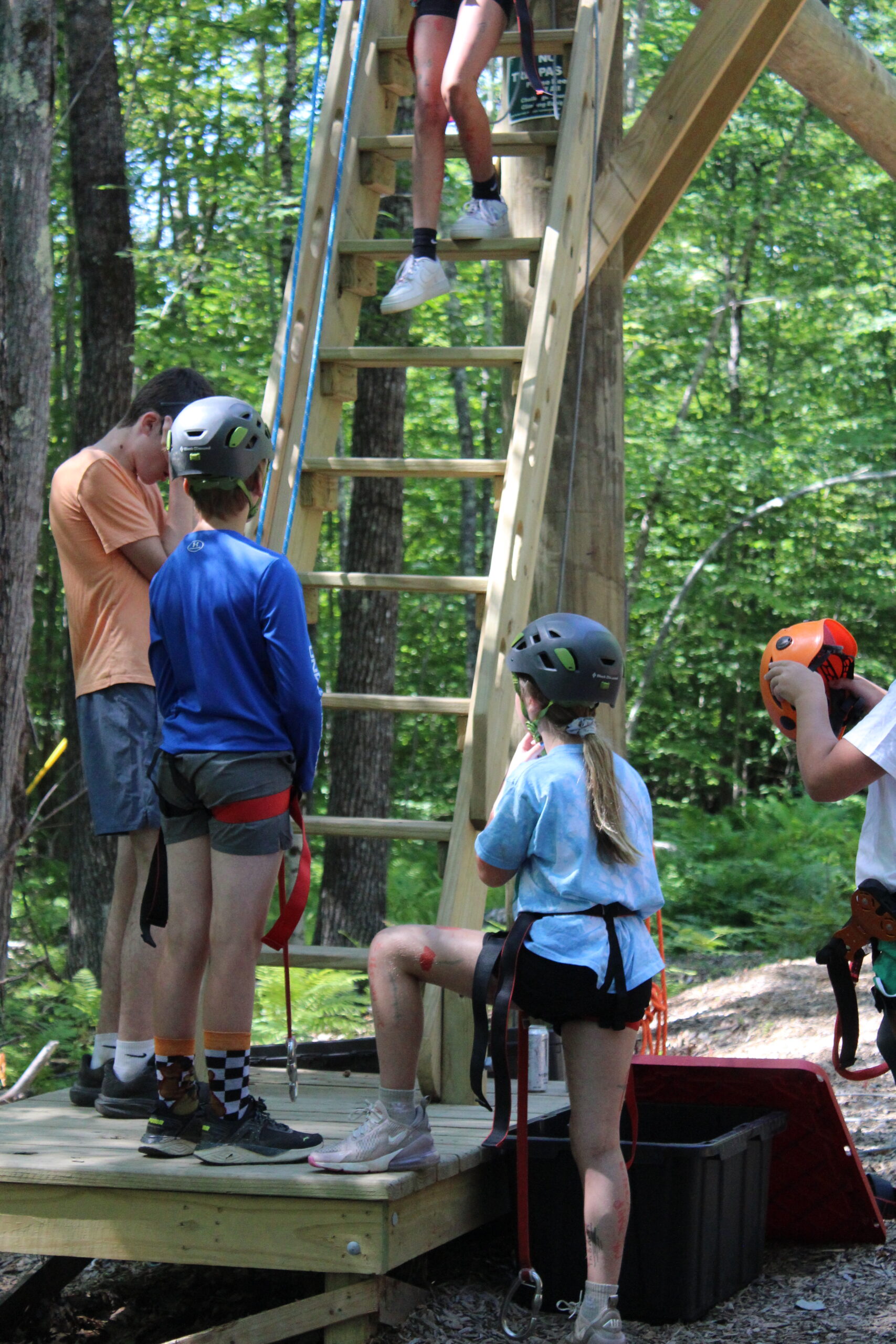 Three youth and a counselor, all wearing protective gear, stand at the bottom of a wooden ladder in the woods. Above them is a large ropes course area.