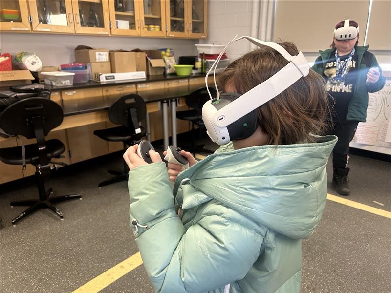 A student in a classroom setting uses a VR headset and holds a wireless controller to interact with a digital environment.