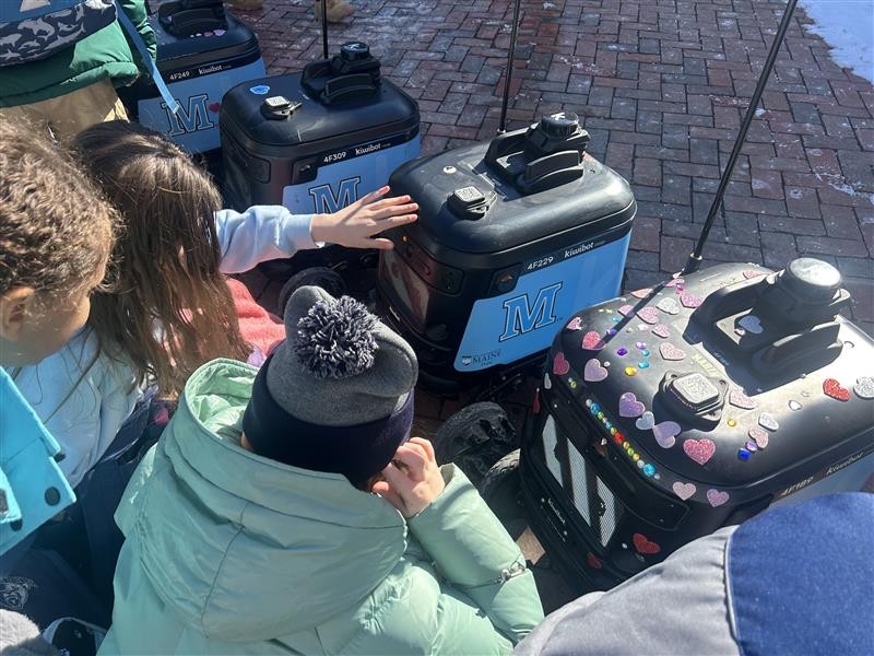 A group of children huddled around several blue and black delivery robots on a brick walkway.
