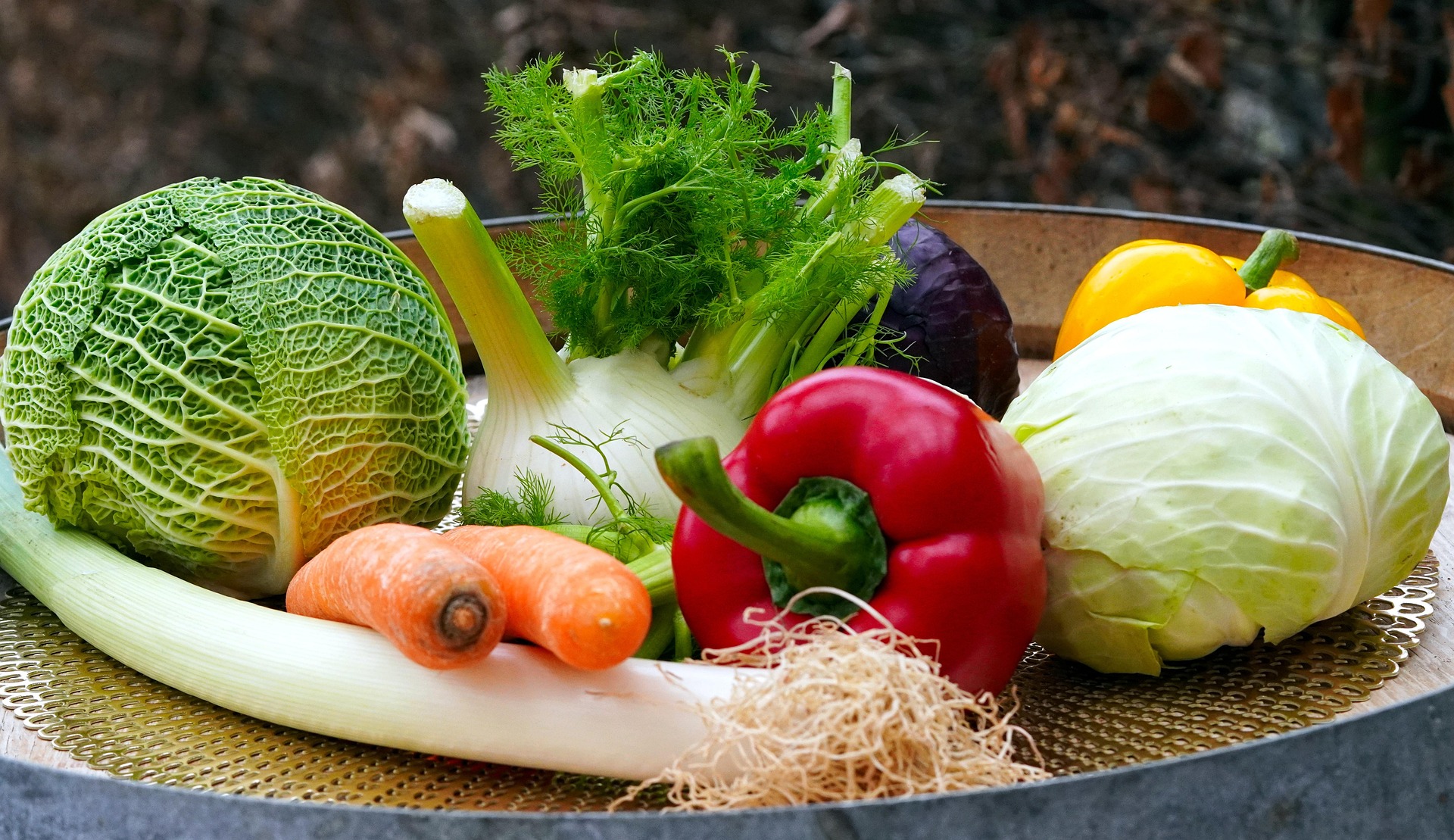 A variety of fresh vegetables on a round tray, including green cabbage, fennel, carrot, leek, red and yellow bell peppers, and a purple vegetable.