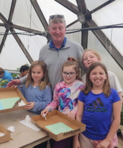 After School Program youth stand smiling with Mary Nash Beaupre Founder, John Beaupre at a recent visit to the greenhouse. In the background are other children and the walls of the greenhouse.