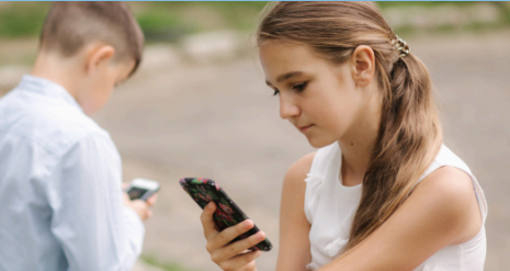 A young boy and girl, facing away from each other, and are both pre-occupied with their mobile devices. They appear to be standing in a parking lot area.