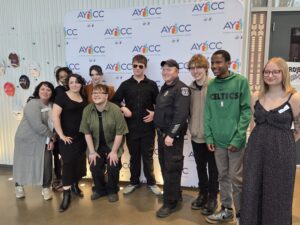 A group of teens, program staff, and Officer Rob Bouley pose for a group photo in front of an AYCC-logoed background.