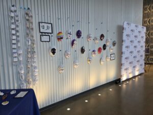 Paper chains hang on a white wall, next to artwork and painted facemasks. They are illuminated by lights on the floor.