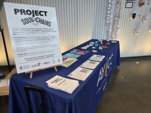 A long table covered with a blue AYCC-logoed tablecloth contains a large sign describing Project Soul Chains. Other paperwork, information, and clay art are at the far end of the table.