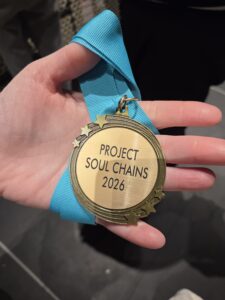 Each teen participant received a gold medalian with "Project Soul Chains 2026" engraved on it. The medallian with a thick light blue ribbon is attached and wrapped around an individual's hand, with the palm facing up.