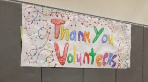 A white banner hangs on a grey wall in a gymnasium. The banner was designed and decorated by youth and says in giant letters, "Thank You Volunteers."